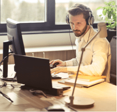 Un hombre con barba y cascos con auriculares trabajando con un portátil con pantalla extra