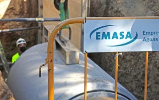 Un operario supervisando la colocación de un trozo de tubería en una zanja mediante una grua, en primer plano tenemos una valla de obra de EMASA